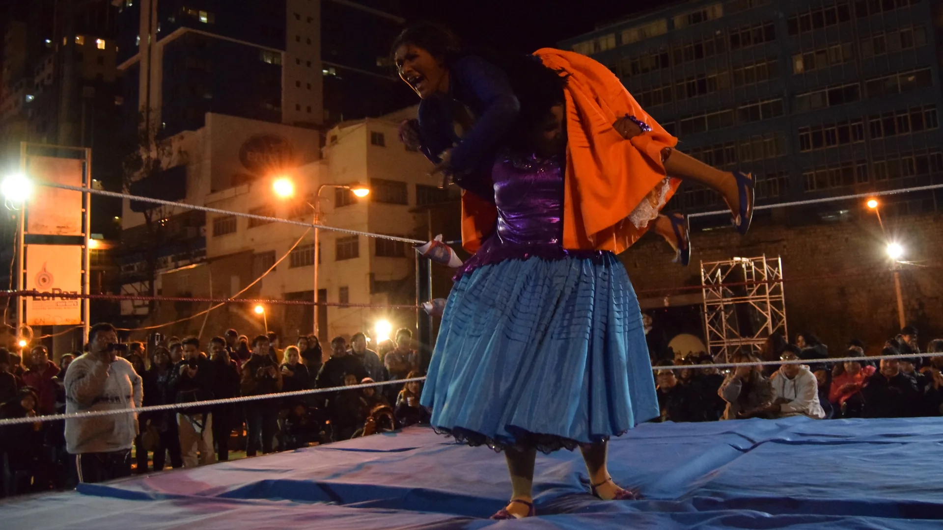 Two cholita wrestlers in a floodlit ring at night, one in a blue pollera lifting the other across her shoulders, with a crowd and city buildings behind the ropes