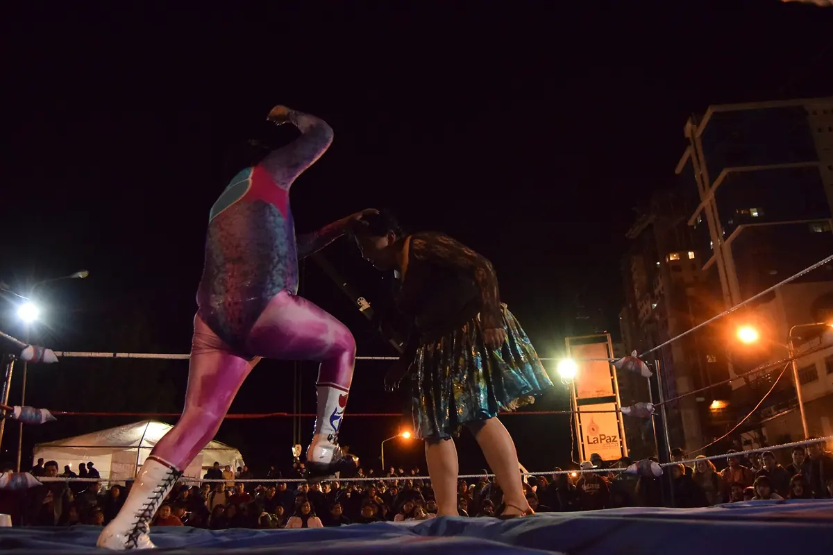 A male wrestler in a pink and grey bodysuit squaring up to a cholita in a green patterned pollera in a floodlit ring at night