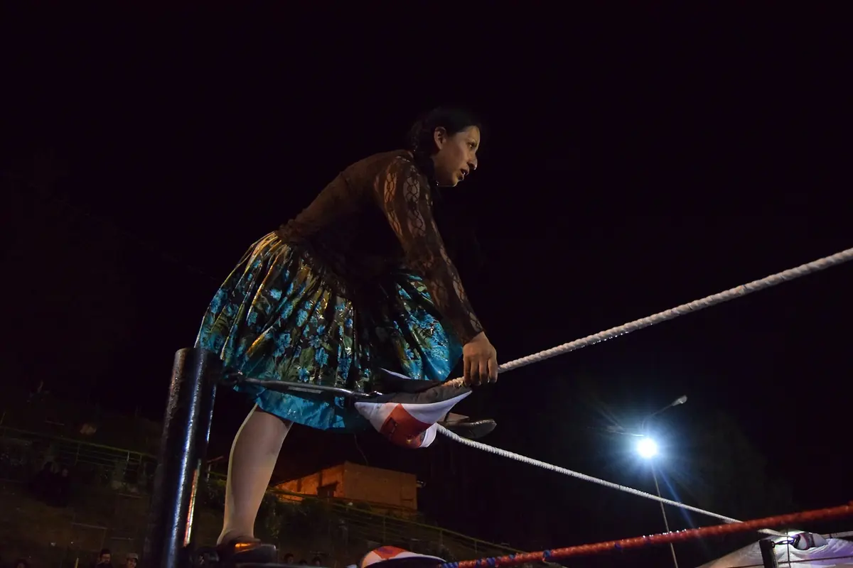 A woman in a dark top and a turquoise and green patterned pollera stepping through the ropes of a ring at night