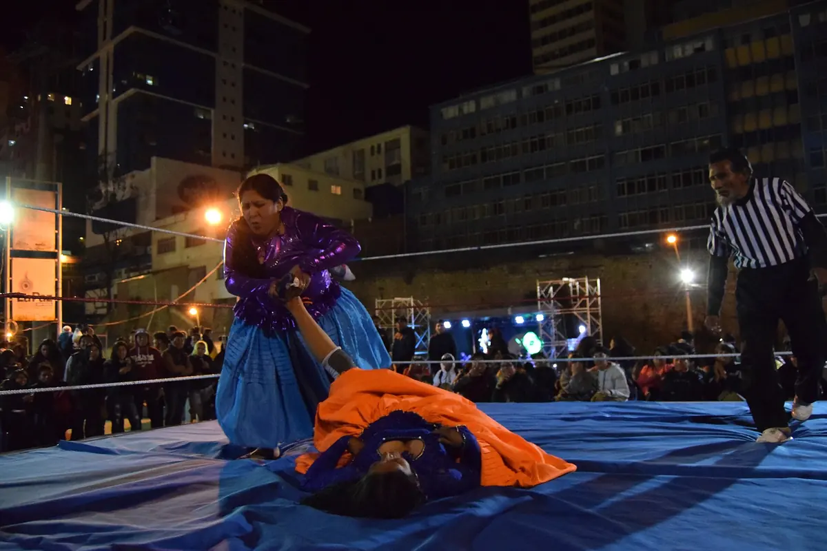 A cholita wrestler in a purple top and blue pollera standing over an opponent lying on the mat in an orange skirt, with a referee in a striped shirt on the right